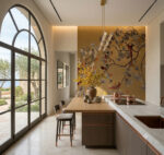 A luxurious modern bedroom with a large gold and ginkgo-patterned wallpaper feature wall, featuring stylized branches, yellow, grey, and red leaves, and exotic birds. The room has a wood-framed bed with white linen, red and ginkgo pillows, and a large arched window with a balcony view of olive trees and the sea. Natural light and a decorative lantern are present.