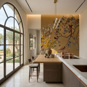 A luxurious modern bedroom with a large gold and ginkgo-patterned wallpaper feature wall, featuring stylized branches, yellow, grey, and red leaves, and exotic birds. The room has a wood-framed bed with white linen, red and ginkgo pillows, and a large arched window with a balcony view of olive trees and the sea. Natural light and a decorative lantern are present.