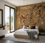 A luxurious modern bedroom with a large gold and ginkgo-patterned wallpaper feature wall, featuring stylized branches, yellow, grey, and red leaves, and exotic birds. The room has a wood-framed bed with white linen, red and ginkgo pillows, and a large arched window with a balcony view of olive trees and the sea. Natural light and a decorative lantern are present.
